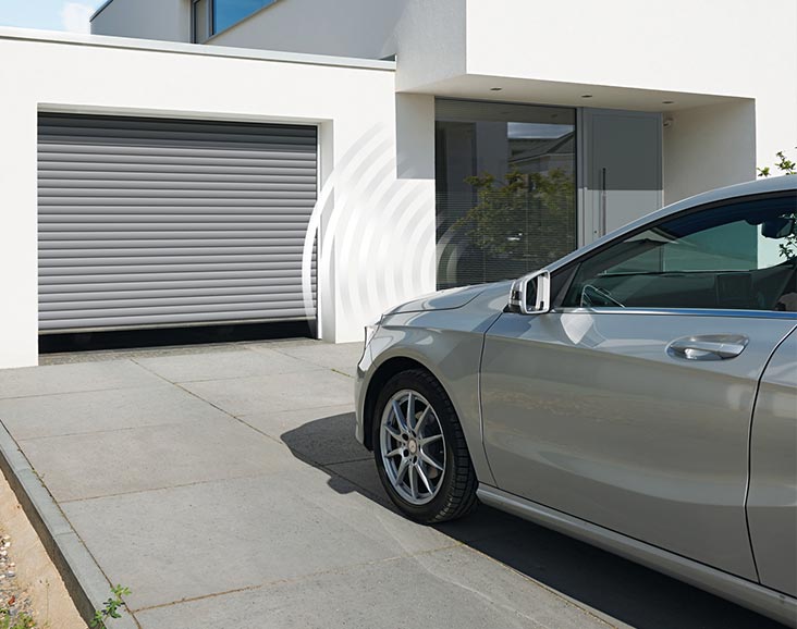 Auto fährt auf elektrische Garage zu, die sich automatisch öffnet, mit Haustür im Hintergrund, zeigt die Bequemlichkeit und Integration unserer Alm-Tor Produkte.