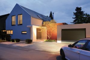 Modernes Haus in einer Wohnsiedlung am Abend mit sich öffnender Garage und beleuchtetem Innenraum, symbolisiert ein herzliches Willkommen zu Hause.